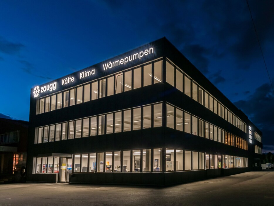Das Firmengebäude der f. zaugg ag in Thun bei Nacht. Sämtliche Räume des Gebäudes sind hell erläuchtet und hinter dem Gebäude erkennt man einen dunkelblauen Himmel. - Ihr Partner für Kühlung, Klimaanlagen und Wärmepumpen.